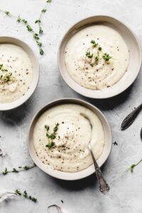 three bowls of creamy cauliflower soup on a grey backdrop in a light bowl decorated with thyme