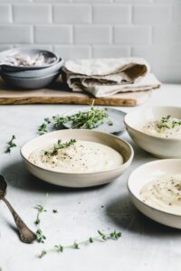 three light brown bowls of creamy vegan cauliflower soup topped with thyme on a grey backdrop with a wooden cutting board in the background together with fresh thyme on a small blue plate and a linen light brown napkin
