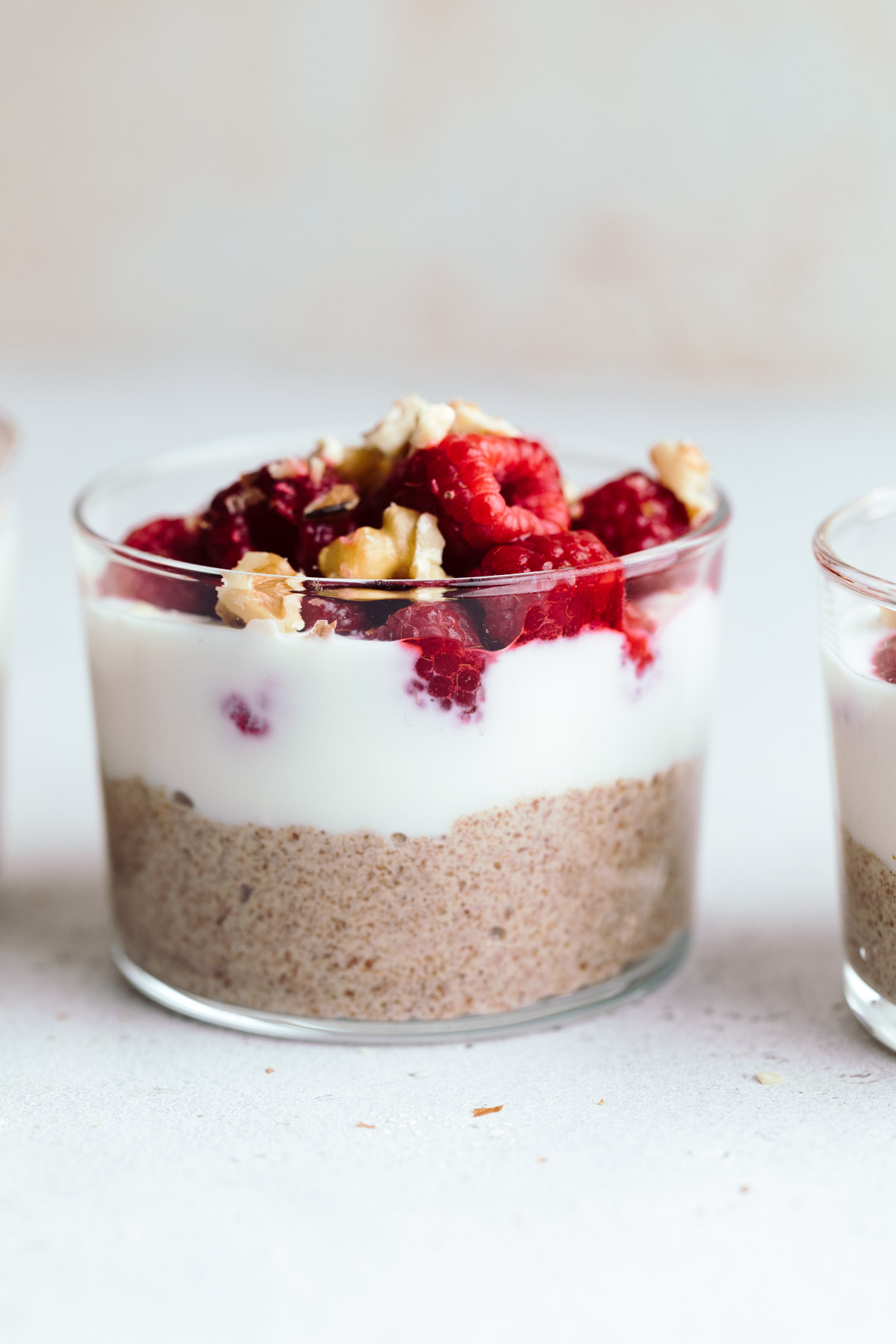 A small glasses layered with brown pudding, then white yogurt and then red raspberries with chopped walnuts on a light grey backdrop.