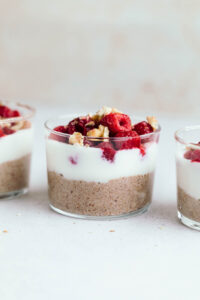Three small glasses in a row layered with brown pudding, then white yogurt and then red raspberries with chopped walnuts on a light grey backdrop.