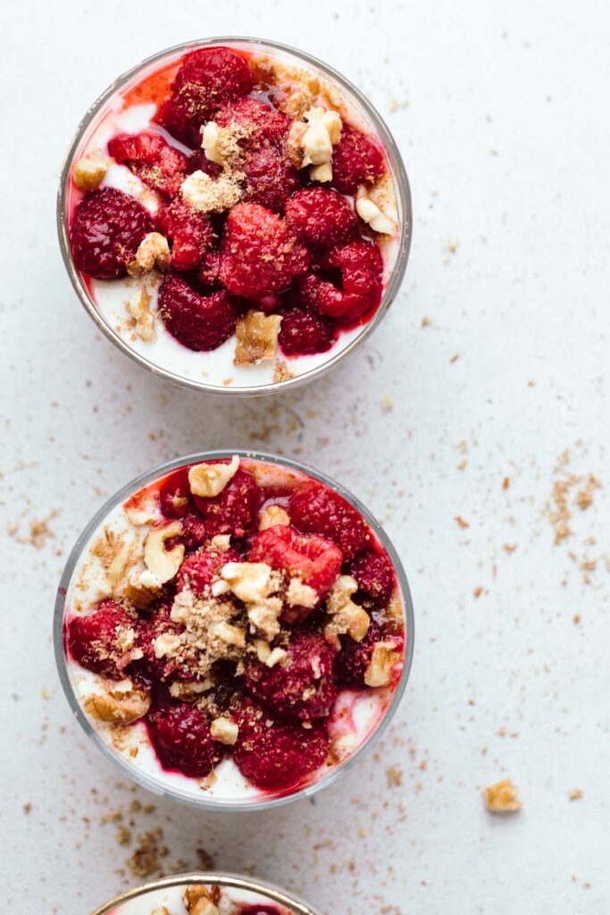 The top view of two glasses with yogurt, raspberries and chopped walnuts on a light grey backdrop and a third glass showing only the edges on the bottom.