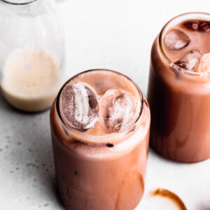two glasses of iced chocolate next to each other with ice floating to the top on a dark grey backdrop with an oat milk bottle in the background
