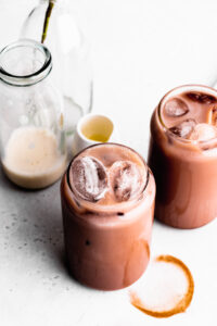 two glasses of iced chocolate next to each other with ice floating to the top on a dark grey backdrop with small bottles in the background
