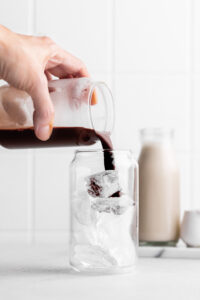 glass with ice cubes and a hand pouring in the chocolate mixture on a white backdrop with a bottle of oat milk in the back