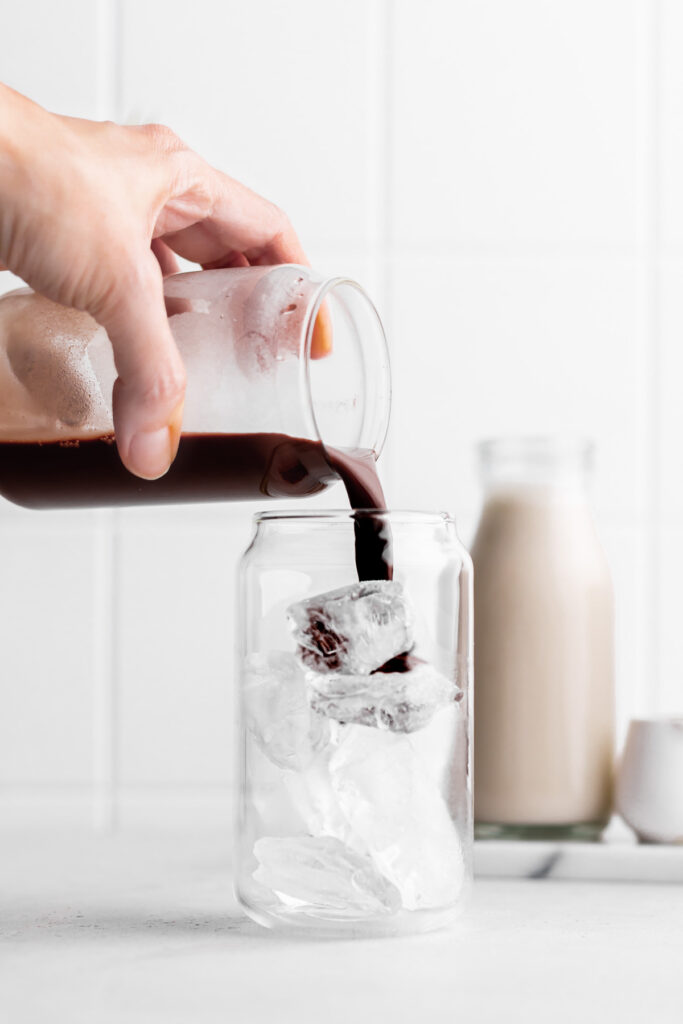 glass with ice cubes and a hand pouring in the chocolate mixture on a white backdrop with a bottle of oat milk in the back