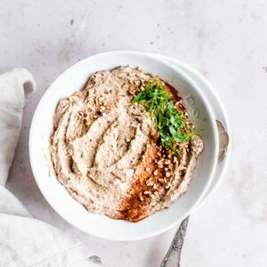 baba ganoush in a light bowl on a plate on a light background with fresh herbs, smoked paprika powder and chopped hazelnuts on top next to a light brown napkin