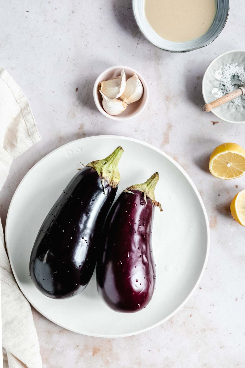 ingredients for baba ganoush on a light backdrop with two eggplants on a plate and tahini in a small bowl with three garlic cloves in a small bowl and half a lemon cut open and salt in pinch bowl with a small wooden spoon