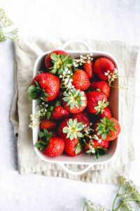 fresh strawberries with stem and little white flowers in a square white dish on a light brown napkin with a light backdrop