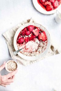 pan roasted strawberries in a light bowl with yogurt and oats on a light backdrop with a light brown napkin and a small pitch bowl with oats and a hand