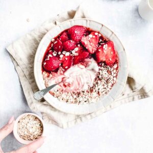 pan roasted strawberries in a light bowl with yogurt and oats on a light backdrop with a light brown napkin and a small pitch bowl with oats and a hand