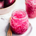 Glass jar with easy pickled red onions on a small blue plate with a wooden fork on a grey backdrop with red onions and another glass jar with pickled red onions in the back.