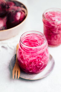Glass jar with easy pickled red onions on a small blue plate with a wooden fork on a grey backdrop with red onions and another glass jar with pickled red onions in the back.