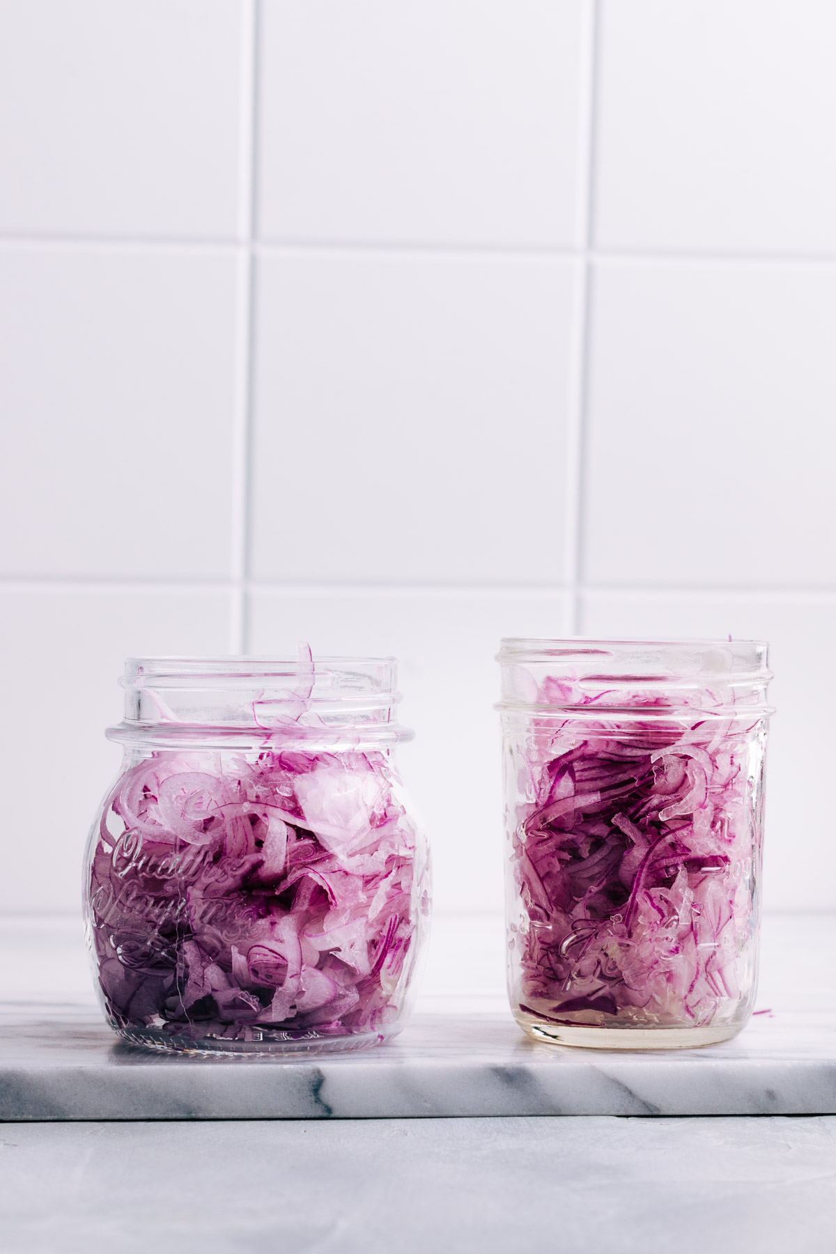 Two glass jars with thinly sliced red onions on a marble cutting board with a white tile backdrop.