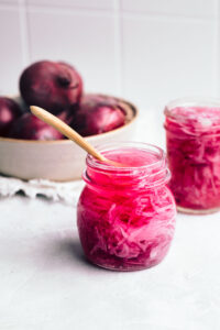 Glass jar with easy pickled red onions on a small blue plate with a wooden fork in it on a grey backdrop with red onions and another glass jar with pickled red onions in the back.