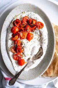 charred tomatoes on yogurt in an oval plate with a silver spoon and herbs on top and pieces of flat bread next to it