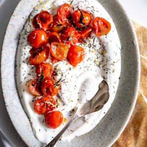 charred tomatoes on yogurt in an oval plate with a silver spoon and herbs on top and pieces of flat bread next to it