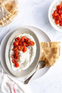 charred tomatoes on yogurt in an oval plate with a silver spoon and herbs on top and pieces of flat bread next to it on a white backdrop with flatbread and more charred tomatoes on yogurt on seperate plates with a white napkin