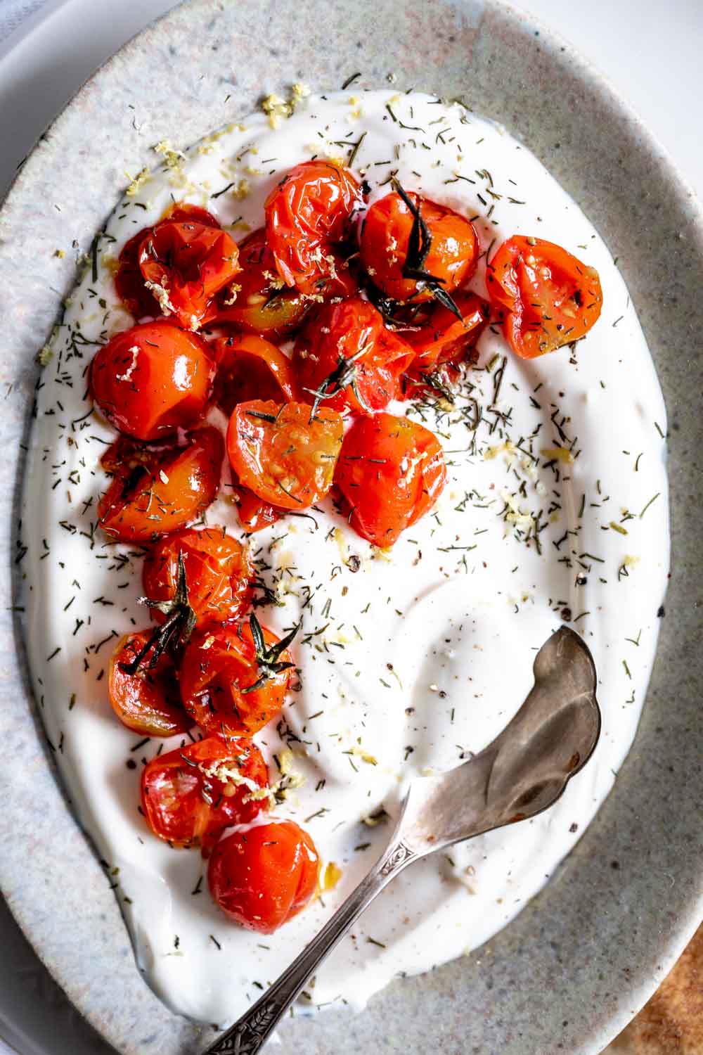 charred tomatoes on yogurt in an oval plate with a silver spoon and herbs on top