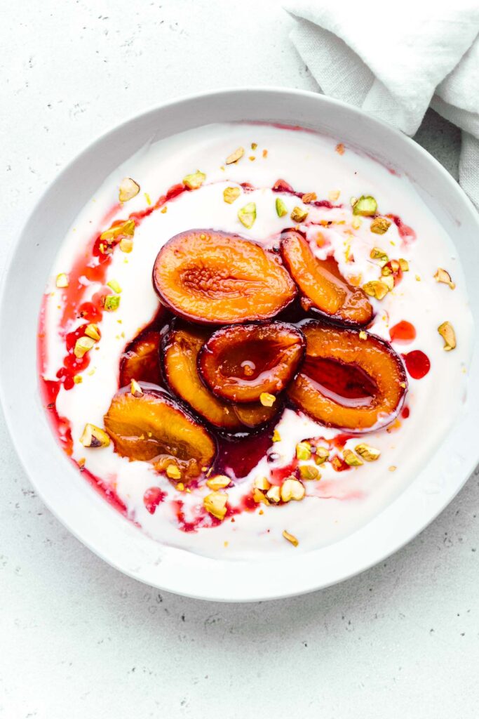 A white bowl on a white backdrop with white yogurt in it topped with red and dark orange glossy stewed plum halves, drizzled with a red syrup and chopped pistachio nuts.