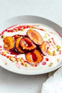 A light grey bowl on a light grey backdrop with white yogurt in it topped with red and dark orange glossy stewed plum halves, drizzled with a red syrup and chopped pistachio nuts.