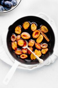 A white skillet with black interior with red and dark yellow stewed plum halves and a cinnamon stick in a glossy liquid and a golden spoon in it on a white napkin on a light backdrop with a bowl with whole plums in the left top corner.