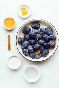 A light grey backdrop with a large bowl with blue plums in it and smaller bowls around it with orange juice, orange zest, water, a cinnamon stick and white sugar.