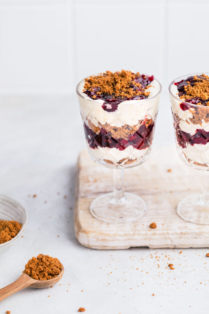 Two tall glasses with vegan parfait on a white wooden board with a wooden spoon next to it with crumbled cookies.