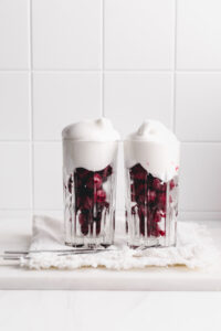 two glasses on a white napkin with cherries and whipped cream on top on a white backdrop and a silver dessert spoon in front