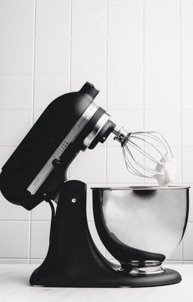 black kitchenaid mixer with aquafaba whipped cream on the whisk with a stainless steel bowl and a white backdrop