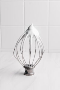 a whisk with whipped aquafaba with a white tile background