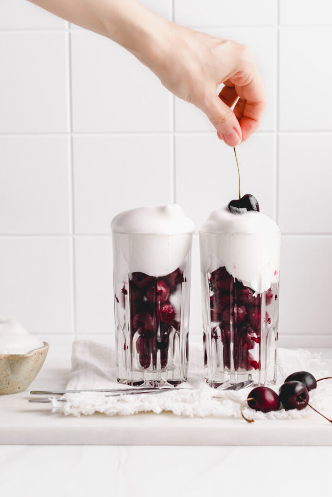 two glasses with cherries and whipped cream on top and a hand dipping one cherry in the whipped cream on a white backdrop with a small bowl of whipped cream next to the glasses