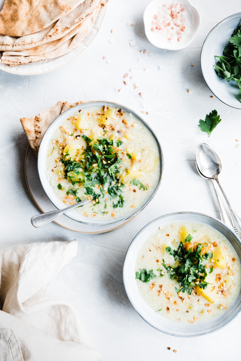 two bowls of cooked creamy potato soup with silver spoons and flatbread on a white background