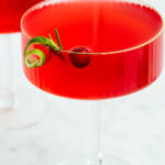 A coupe cocktail glass with a red drink decorated with a small rosemary twig a cranberry and a lime zest curl on a white marble backdrop in front of another glass with a red drink.
