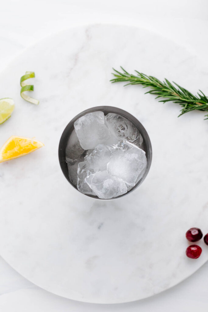 A top view of a cocktail shaker on a round marble backdrop filled with ice cubes next to a few cranberries a rosemary twig and a lime zest curl.