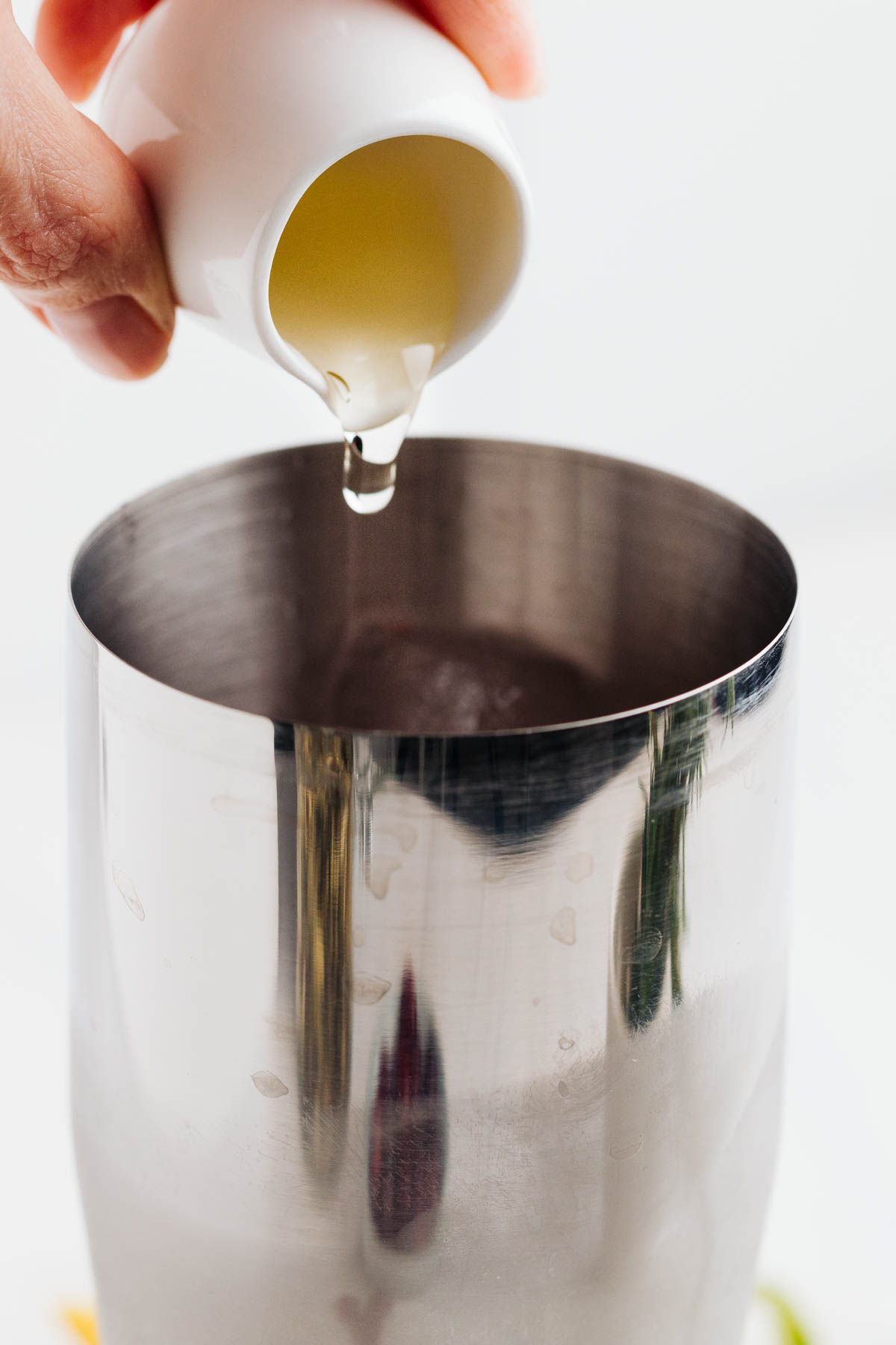 A silver colored cocktail shaker with a light colored syrup liquid being poured in.