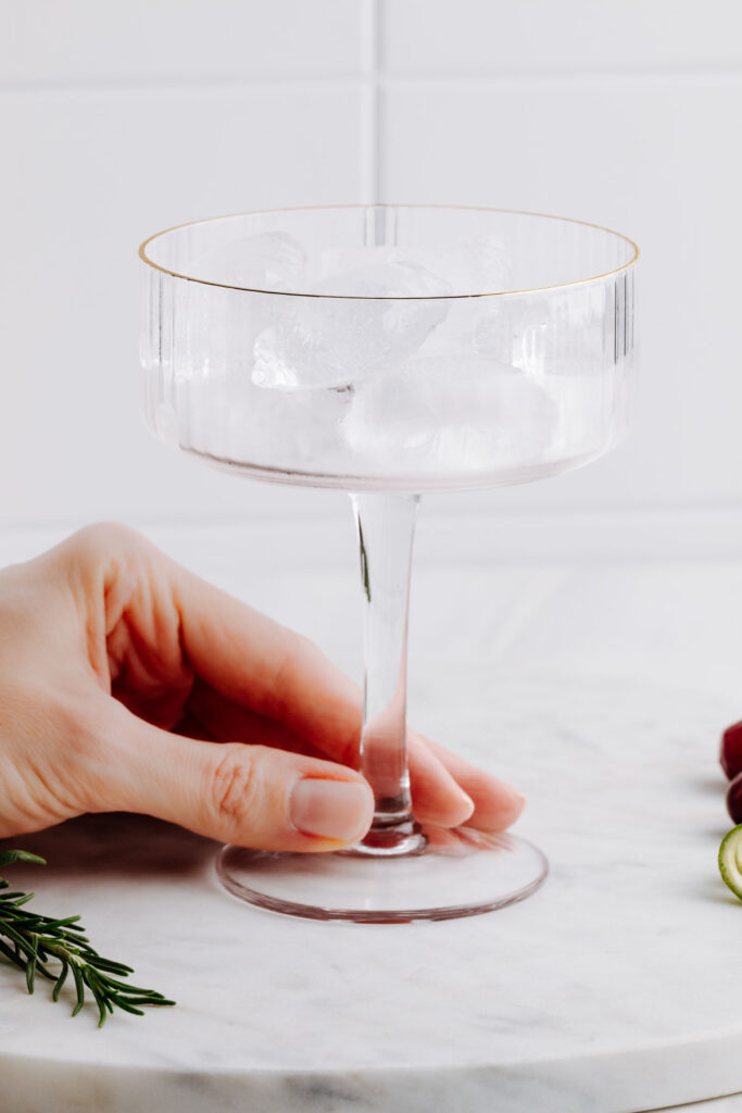 A coupe cocktail glass on a white marble cutting board in front of a white tiled backdrop filled with ice cubes and a hand holding the stem of the glass.