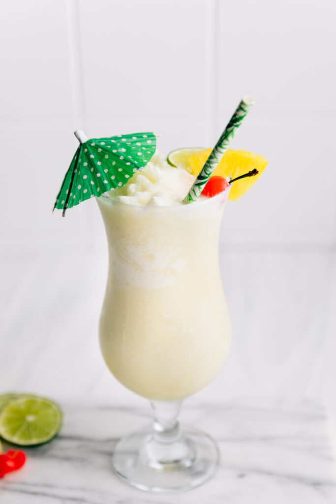 Piña Colada Mocktail in a tall hurricane glass decorated with a green paper umbrella, a pineapple chunk, lime, a cherry and a green paper straw on a marble cutting board on a white backdrop with slices of lime, and cherries next to the glass