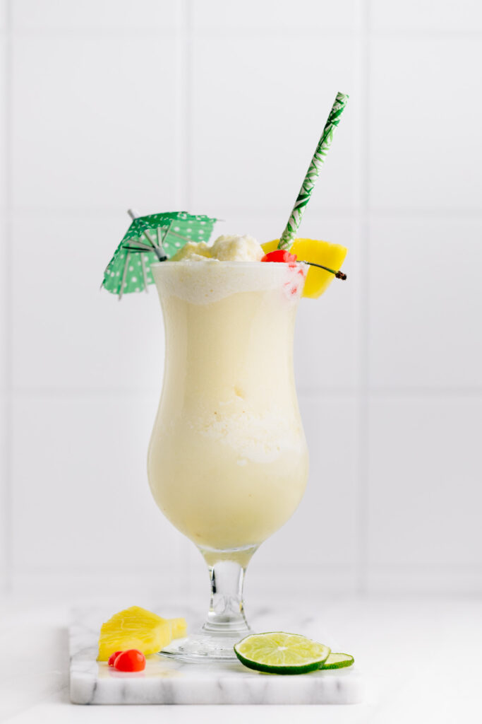 Piña Colada Mocktail in a tall hurricane glass decorated with a green paper umbrella, a pineapple chunk, a cherry and a green paper straw on a marble cutting board on a white backdrop with slices of lime, pineapple and cherries next to the glass