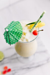 Piña Colada Mocktail in a tall hurricane glass from the top decorated with a green paper umbrella, a pineapple chunk, a cherry, a slice of lime and a green paper straw on a marble cutting board on a white backdrop