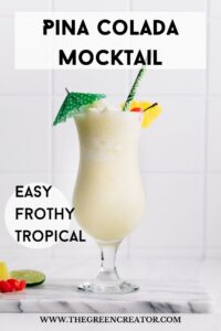 Piña Colada Mocktail in a tall hurricane glass decorated with a green paper umbrella, a pineapple chunk, a cherry and a green paper straw on a marble cutting board on a white backdrop with text over it for Pinterest