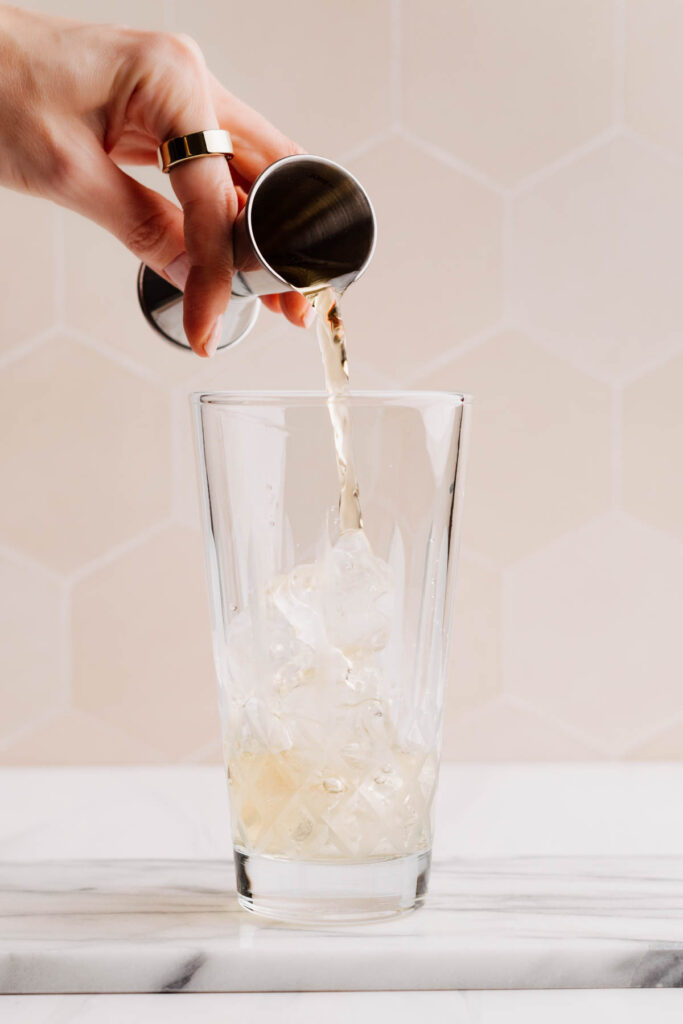A glass cocktail shaker with ice cubes and whiskey being poured over the ice with a jigger.
