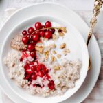 red currant oat porridge topped with red currant and chopped walnuts in a white bowl on a white plate on a light wooden cutting board with a golden spoon in the porridge