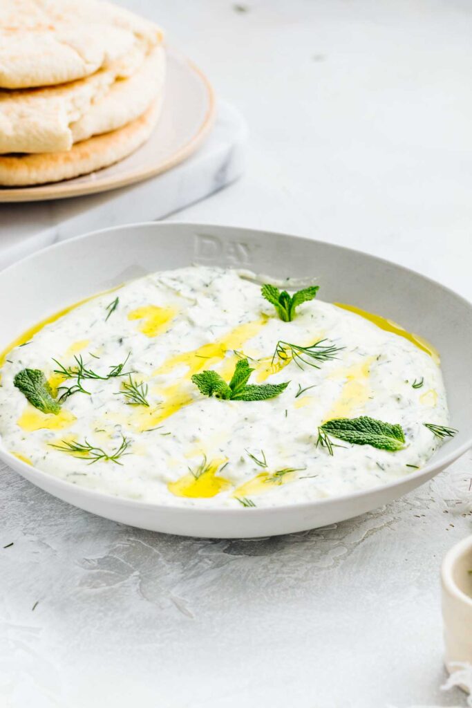 A bowl on a light grey backdrop with a thick dip in it garnished with olive oil, dill and mint leaves in front of a stack of pita breads on a marble cutting board.