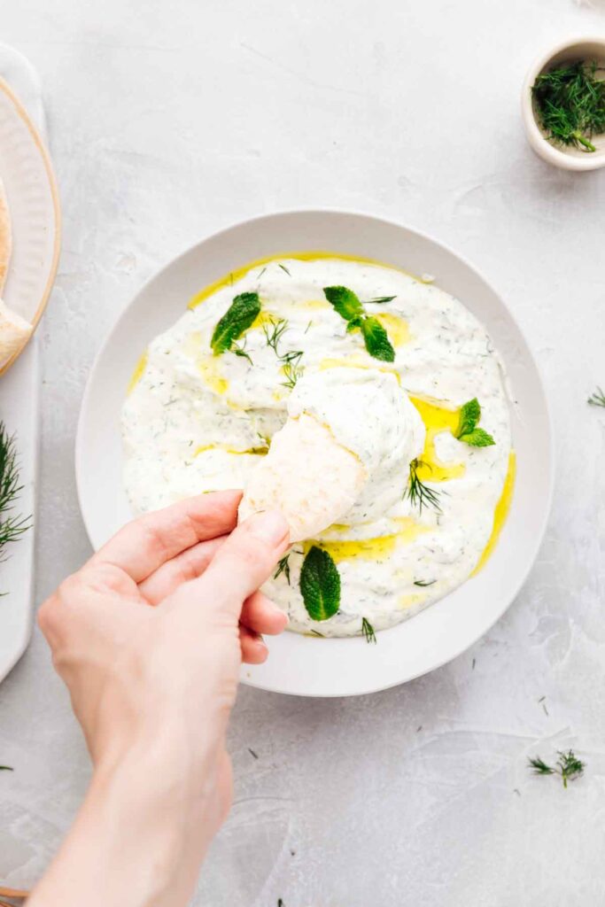 A bowl on a light grey backdrop with a thick dip in it garnished with olive oil, dill and mint leaves and a hand holding a piece of pita bread dipping it in the sauce.