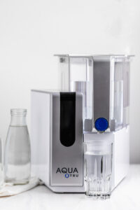 AquaTru water filter on a white surface next to a glass water bottle and a full water glass