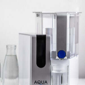 AquaTru water filter on a white surface next to a glass water bottle and a full water glass