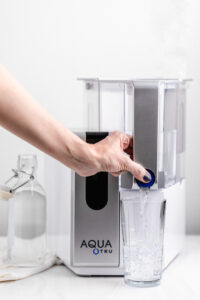 AquaTru water filter on a white surface next to a glass water bottle and a hand pressing the dispense button filling in a glass with water