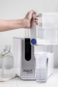 AquaTru water filter on a white surface next to a glass water bottle and a hand lifting the front water tank