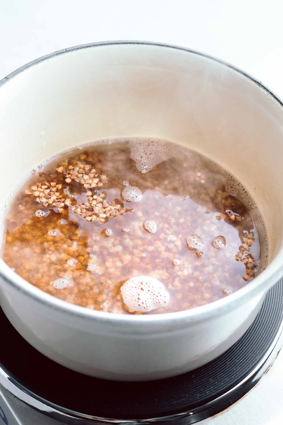 A light colored pot on a small stove with water and roasted buckwheat in it and steam coming of the water.