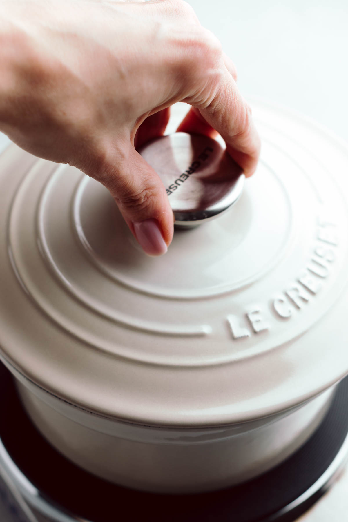 A light colored pot on a stove with a lid and a hand on top of the lid.